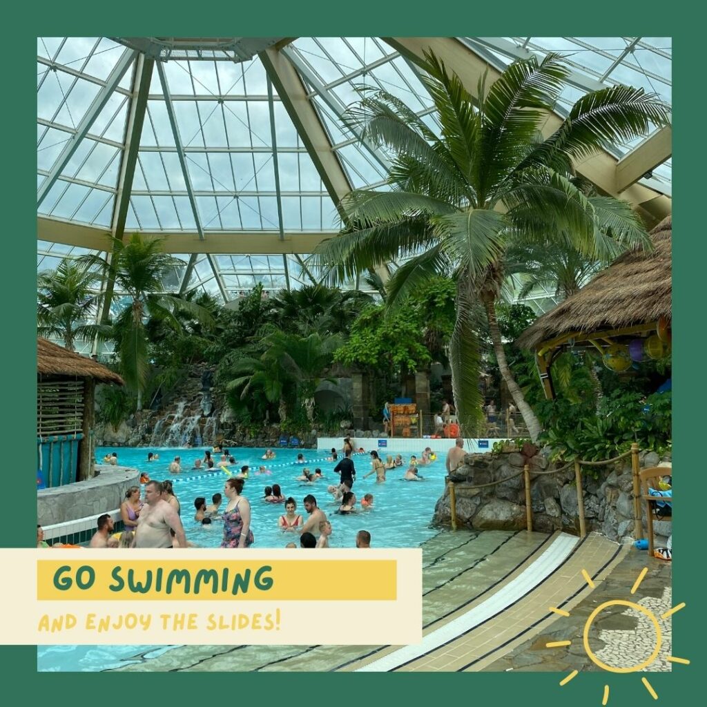 Text: Go swimming (and enjoy the slides!). Image: inside the Center Parcs Subtropical Paradise, people swimming in the pool, palm trees around.