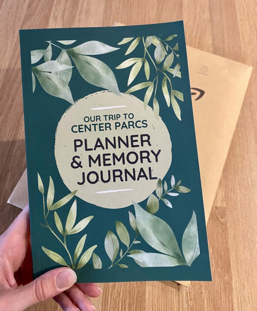 Green book with leaves being held above Amazon packaging on a wooden background. The title of the book says: "Our trip to Center Parcs: Planner & Memory Journal"